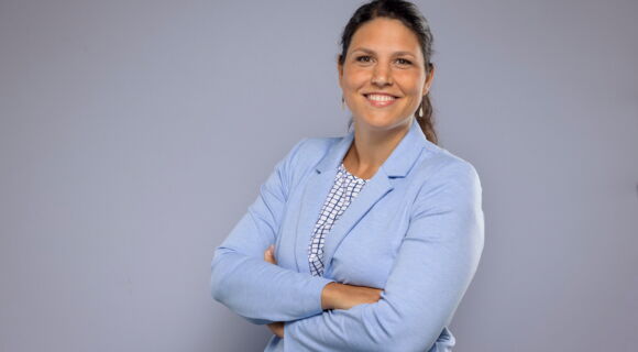 Sarah Karge, eine Frau mit brauen, langen Haaren, steht mit verschränkten Armen vor einem neutralen Hintergrund. Sie trägt einen hellblauen Blazer über einer gemusterten Bluse. Sie lächelt offen in die Kamera.
