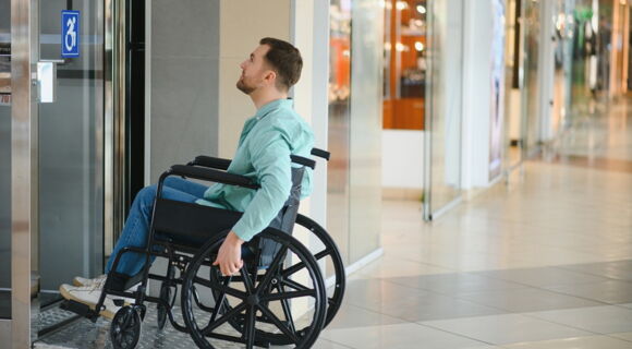 Ein Rollstuhlfahrer vor einer Tür in einem Einkaufszentrum. Auf der Glastür ist ein Aufkleber mit Rollstuhl angebracht.