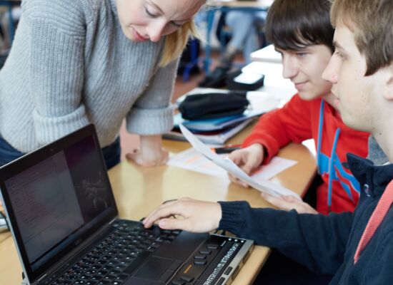 In einem Klassenraum beugt sich eine junge Lehrerin über den Laptop mit Braillezeile eines Schülers. Andere Jugendliche sitzen mit im Raum.