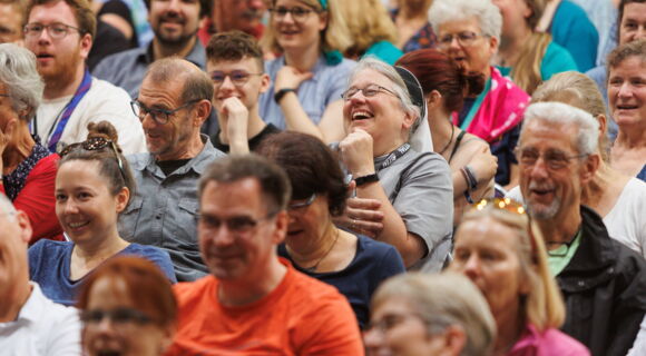 Bild eines Publikums beim Kirchentag 2023 in Nürnberg: Lachende und lächelnde Menschen verschiedenen Alters blicken in eine Richtung. Einige tragen kurzärmelige T-Shirts.