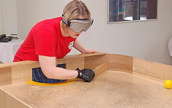 Eine Frau steht an einem Showdown-Tisch mit hohem Rand und Toren. Sie schlägt mit einem flachen, kleinen Schläger den Ball zurück. Ihre Augen sind von einer Brille bedeckt.