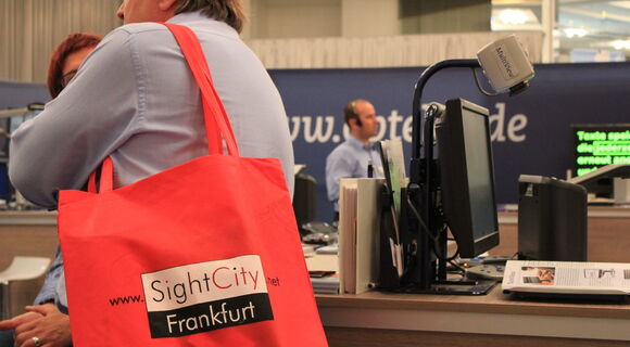 Ein Bild aus dem Messegeschehen. Im Vordergrund leuchtet eine rote Tasche, auf der "SightCity Frankfurt" steht.