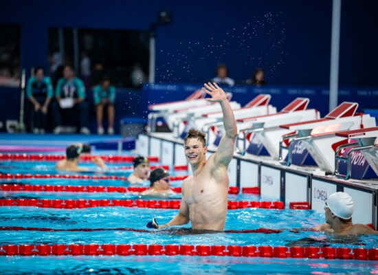 Taliso Engel winkt mit einem breiten Lachen aus dem Wasser in einem Schwimmbad. Er ist ein sehr muskulöser junger Mann. Neben ihm sind andere Schwimmer mit Badekappen.