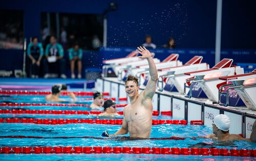 Taliso Engel winkt mit einem breiten Lachen aus dem Wasser in einem Schwimmbad. Er ist ein sehr muskulöser junger Mann. Neben ihm sind andere Schwimmer mit Badekappen.