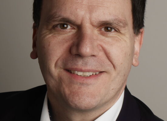 Prof. Dr. Thomas Ach blickt lächelnd in die Kamera. Er hat dunkles kurzes volles Haar mit geradem Ansatz. Er trägt ein schwarzes Sakko und ein weißes Hemd.