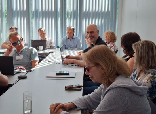Beim Workshop „Überregionale Kooperation“: Teilnehmerinnen und Teilnehmer sitzen an Tischen. Vor manchen liegt ein Smartphone, andere haben ihr Laptop aufgeklappt. Auch Workshop-Leiter Reiner Delgado bedient ein Laptop, während er lächelnd Richtung Kamera