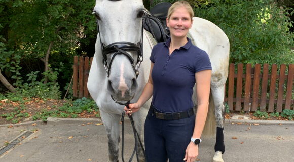 Helen Vogt steht mit ihrer Schimmelstute Janita auf einem Reitplatz, der von Büschen und Bäumen umgeben ist. Sie hat langes blondes, zu einem Pferdeschwanz gebundenes Haar und trägt ein kurzärmeliges Polohemd. Janita trägt einen Sattel und Zaumzeug.