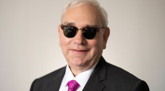 Portrait von Hans-Werner Lange. Er hat kurze, weiße Haare und ein volles Gesicht. Er trägt ein weißes Hemd, darüber ein dunkles Jackett, dazu eine purpurfarbene Krawatte und eine dunkel getönte Brille. Er schaut lächelnd in die Kamera.