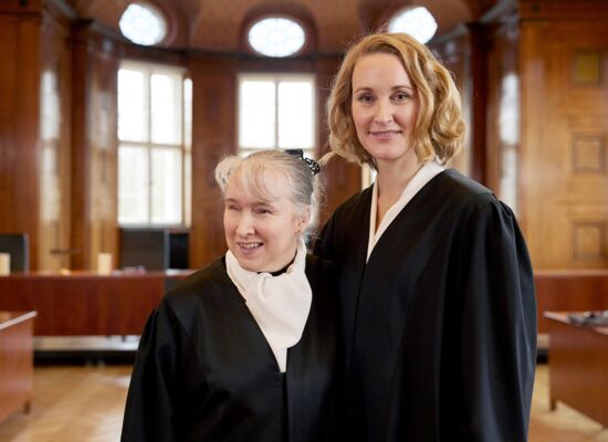 Pamela Pabst und Christina Athenstädt, beide in schwarzer Robe, stehen in einem  Gerichtssaal, Pamela Pabst trägt ihr helles Haar hochgesteckt.