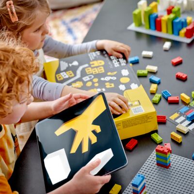Zwei Kinder sitzen an einem Tisch. Vor sich liegt ein Karton mit Braille-Bricks, darum herum Legosteine.