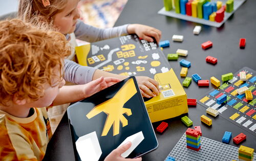 Zwei Kinder sitzen an einem Tisch. Vor sich liegt ein Karton mit Braille-Bricks, darum herum Legosteine.
