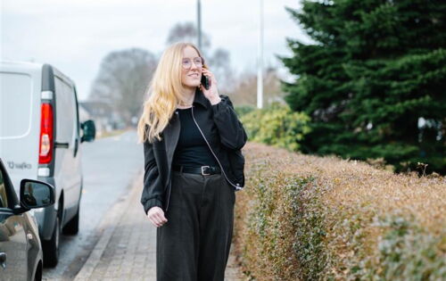 Claire Horsbrugh steht telefonierend an einer Straße. Sie hat langes blondes Haar und trägt eine schwarze Jacke und ein schwarzes Oberteil. Im Hintergrund sind Bäume und Büsche zu sehen.