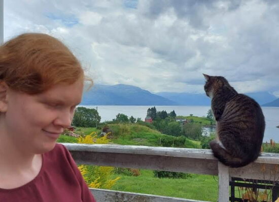 Solveig Marie Oma steht links im Bild und blickt nach unten. Rechts sitzt eine getigerte Katze auf einem Geländer. Im Hintergrund sind ein Fjord, grüne Landschaft, Berge und ein bewölkter Himmel zu sehen.
