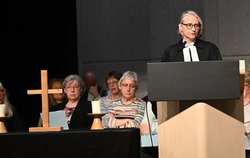 Die Oberkirchenrätin Annette Noller steht im schwarzen Talar am Rednerpult. Sie hat glatte, graue Haare und eine schwarze Brille Auf einem Altar links im Bild stehen ein Holzkreuz und eine weiße Kerze. Dahinter in mehreren Reihen die Mitglieder des Chors.