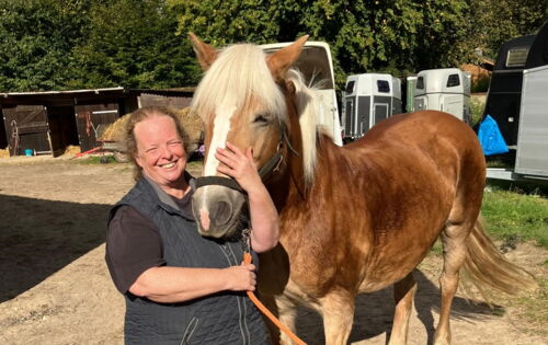 Linda Pröve steht bei einem Haflingerpony, dessen Kopf sie mit einer Hand umfasst. Ihr langes lockiges Haar trägt sie zusammengebunden. Im Hintergrund sind Bäume und Pferdeanhänger zu sehen.