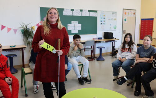 Mandy Kugel steht in einer Schulklasse in einem Stuhlkreis; ein Teil der Kinder ist zu sehen. Sie hat lange blonde Haare und trägt einen warmen Pullover. Mit der linken Hand hält sie ihren Langstock, mit beiden Händen zeigt sie eine Armbinde mit drei Punk