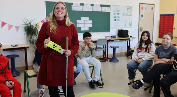 Mandy Kugel steht in einer Schulklasse in einem Stuhlkreis; ein Teil der Kinder ist zu sehen. Sie hat lange blonde Haare und trägt einen warmen Pullover. Mit der linken Hand hält sie ihren Langstock, mit beiden Händen zeigt sie eine Armbinde mit drei Punk