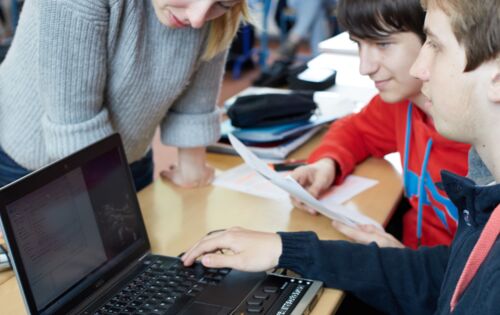 In einem Klassenraum beugt sich eine junge Lehrerin über den Laptop mit Braillezeile eines Schülers. Andere Jugendliche sitzen mit im Raum.