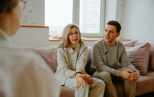 Auf einer Couch sitzen eine Frau und ein Mann, beide etwa Anfang 30. Die Frau hat lange helle Haare, trägt eine Brille und Kreolen. Sie erzählt etwas, er sieht sie an. Vor ihnen sitzt ein Berater, von dem nur der Oberkörper von schräg hinten zu sehen ist.