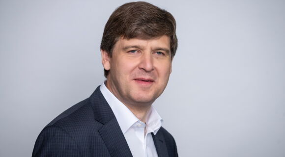 Thomas Krämer hat volles, dunkleres Haar mit einem Seitenscheitel. Er trägt ein weißes Hemd und ein dunkles Sakko.