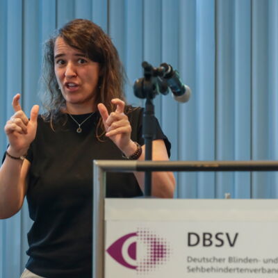 Heike Heubach an einem Stehpult. Sie hat langes welliges Haar. Sie gebärdet mit den Händen. Daumen und Zeigefinger zeigen jeweils einen kleinen Abstand an.
