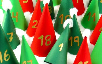 Ein Adventskalender, der aus kleinen bunten, zylindrisch geformten Papphütchen besteht. Darauf ist jeweils eine Zahl zu sehen. Unter jedem Hütchen liegt, wie an einem hochgeklappten zu erkennen ist, eine Leckerei.