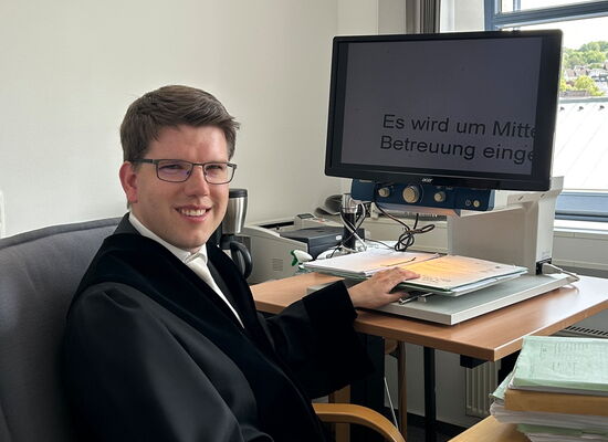 André Stahl sitzt in Richterrobe am Schreibtisch seines Büros. Er hat kurzes dunkleres Haar und trägt eine Brille. Sein Monitor zeigt einen stark vergrößerten Text an.