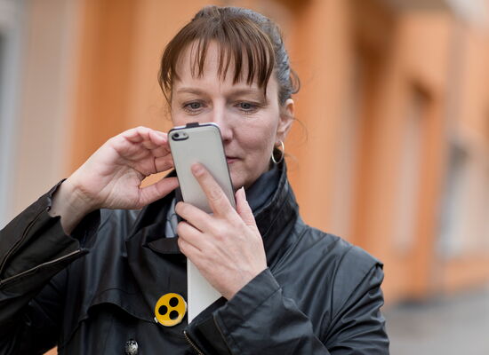Eine Frau mit Pony-Haarschnitt schaut konzentriert auf ihr Smartphone. An ihrer schwarzen Jacke befindet sich ein gelber Anstecker mit drei Punkten.
