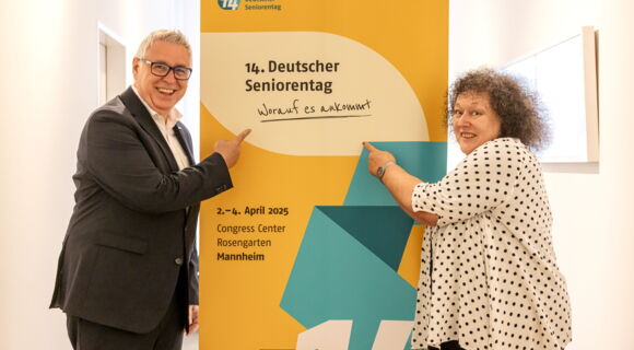 Ein Mann und eine Frau stehen neben einem Aufsteller vom 14. Deutschen Seniorentag und zeigen mit dem Finger darauf. Sie haben die Augen weit geöffnet und lachen. Er trägt einen dunklen Anzug, sie trägt eine gepunktete Bluse und hat lockige, graue Haare.