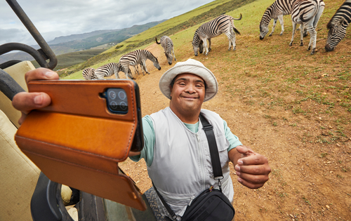 Ein Mann mit gebräunter Haut und einem weißen Hut steht vor ein paar Zebras. Er lächelt. In seiner Hand hält ein Smartphone vor sich als würde er ein Selfie machen. Neben ihm steht ein Geländewagen.