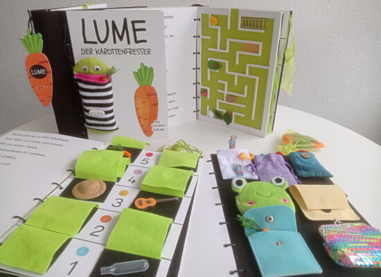 Interaktives Tastbuch „Lume der Karottenfresser“ mit Stofffiguren, Taschen, Labyrinth und verschiedenen taktilen Spielelementen auf mehreren geöffneten Buchseiten.