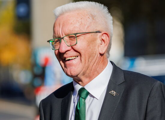 Winfried Kretschmann lachend an einem sonnigen Tag. Er trägt eine grüngerahmte Brille zur grünen Krawatte. Er ist ein Mann Ende 70 mir weißen Haaren.
