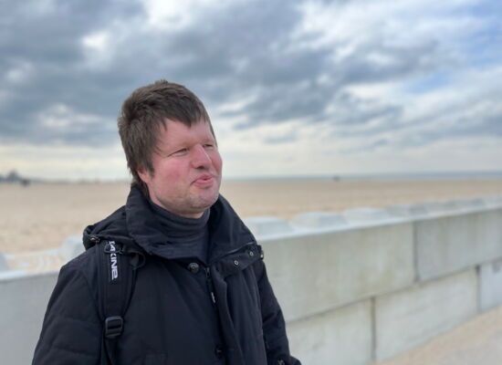 Marco Mers in dunkler Jacke an einem weitläufigen Ostseestrand. Er hat kurzes Haar und lächelt. Der Himmel ist wolkenverhangen.