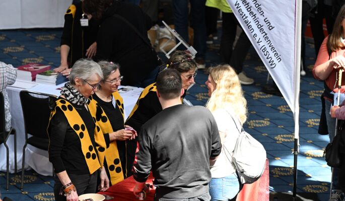 An einem Stand reden fünf Menschen miteinander. Ein Banner markiert den Stand des Blinden- und Sehbehindertenverein Westfahlen e.V. (BSVW). Die Frauen hinter dem Stand tragen gelbe Schärpen mit drei schwarzen Punkten. Sie haben Blumen in den Haaren. Davor