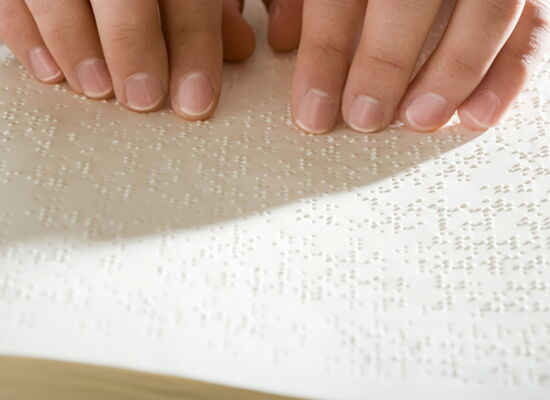 Zwei manikürte Hände lesen Brailleschrift auf weißen Seiten.