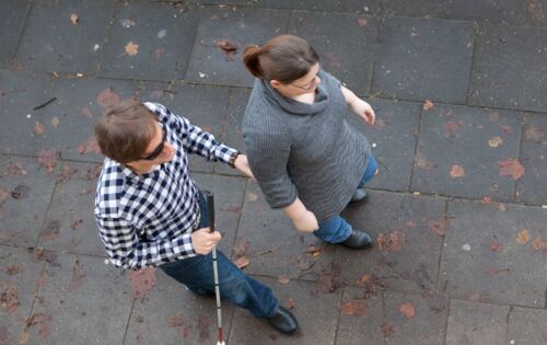 Zwei Menschen gehen eine Straße entlang. Von oben ist zu erkennen, dass sie etwas versetzt hintereinandergehen. Dabei berührt der hintere Mann den Ellenbogen der vorne gehenden Frau. Beide tragen Jeans und grau/schwarze Oberteile.