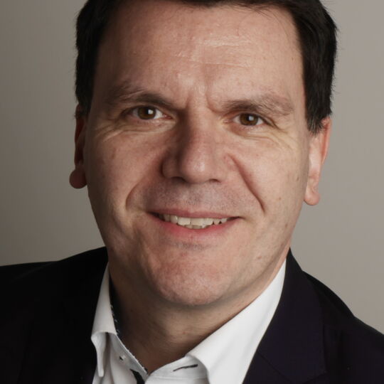 Prof. Dr. Thomas Ach blickt lächelnd in die Kamera. Er hat dunkles kurzes volles Haar mit geradem Ansatz. Er trägt ein schwarzes Sakko und ein weißes Hemd.