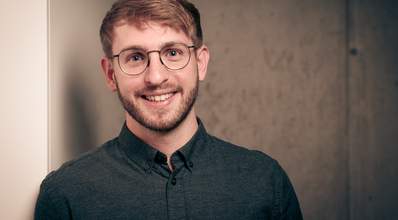 Fabian Rombach lächelt in die Kamera.  Er hat kurzes Haar, trägt eine Brille,  einen kurzen Bart und ein dunkles Hemd.