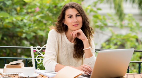 Eine Frau mit langen braunen Haaren sitzt draußen an einem Tisch. Im Hintergrund sind verschwommen Bäume zu sehen. Vor ihr liegen mehrere Bücher und ein Laptop. Die Hand unter dem Kinn haltend, blickt sie versonnen in die Kamera.