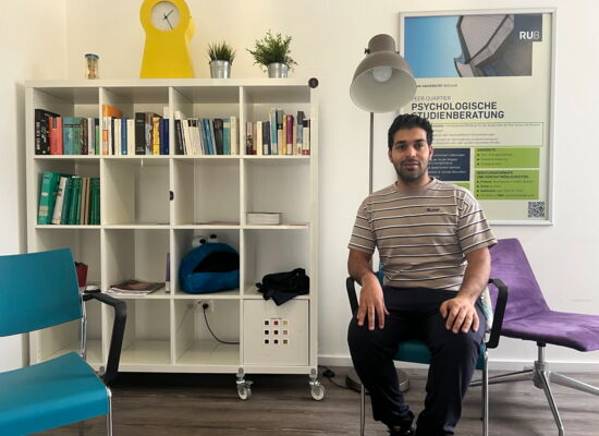 Saiman Mehra sitzt im Raum der Psychologischen Studienberatung. Er hat dunkles Haar und einen Vollbart. Hinter ihm an der Wand steht ein weißes Regal mit quadratischen Fächern, gefüllt mit Büchern und anderen Dingen.