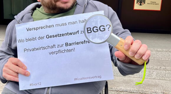 Ein Mann mit kurzen blonden Haaren hält eine Lupe und ein Schild in den Händen. Darauf steht: „Versprechen muss man halten. Wo bleibt der Gesetzentwurf zum BGG? Privatwirtschaft zur Barrierefreiheit verpflichten!“