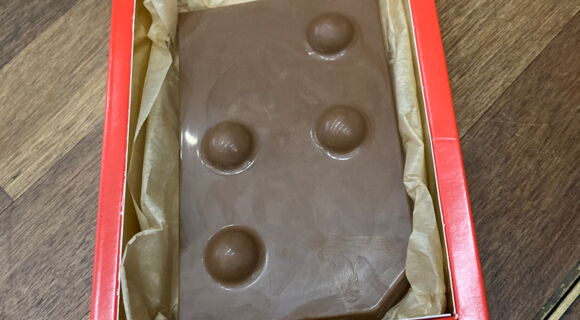 In einer roten Schachtel liegt eine Tafel Schokolade. Darauf ist der Buchstabe "T" in Braille abgedruckt.