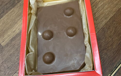 In einer roten Schachtel liegt eine Tafel Schokolade. Darauf ist der Buchstabe "T" in Braille abgedruckt.