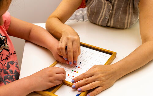 Die Hände eines Mädchens und einer Lehrerin an einem Braille-Board zum Lernen des Alphabets. Sie ertasten die Erhebungen. Beide tragen gemusterte Oberteile und haben lackierte Fingernägel.