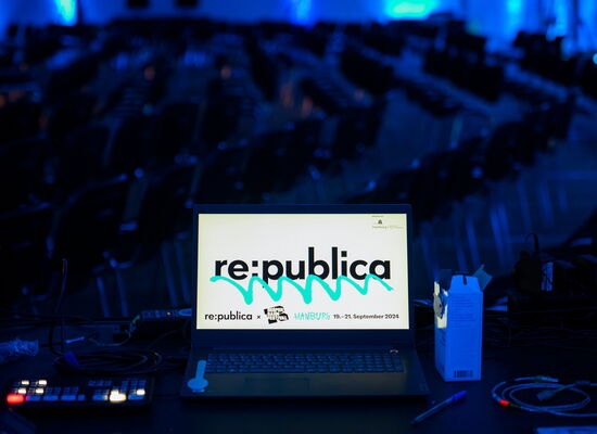 Ein Laptop auf einem Technik-Tisch zeigt das Logo der „re:publica". Im Hintergrund sind viele leere Stuhlreihen in einem blauen Licht beleuchteten Raum zu sehen. Kabel und Technikgeräte liegen auf dem Tisc