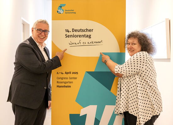 Ein Mann und eine Frau stehen neben einem Aufsteller vom 14. Deutschen Seniorentag und zeigen mit dem Finger darauf. Sie haben die Augen weit geöffnet und lachen. Er trägt einen dunklen Anzug, sie trägt eine gepunktete Bluse und hat lockige, graue Haare.