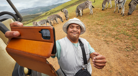 Ein Mann mit gebräunter Haut und einem weißen Hut steht vor ein paar Zebras. Er lächelt. In seiner Hand hält ein Smartphone vor sich als würde er ein Selfie machen. Neben ihm steht ein Geländewagen.