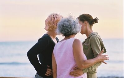 Drei Frauen stehen kreisförmig zusammen und besprechen etwas. Ihre Gesichter sind nicht zu sehen. Zwei von ihnen halten sich gegenseitig unterstützend am Arm. Zwei der Frauen haben nackenlanges blondes lockiges Haar, die dritte hat dunkleres Haar.