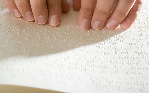 Zwei manikürte Hände lesen Brailleschrift auf weißen Seiten.