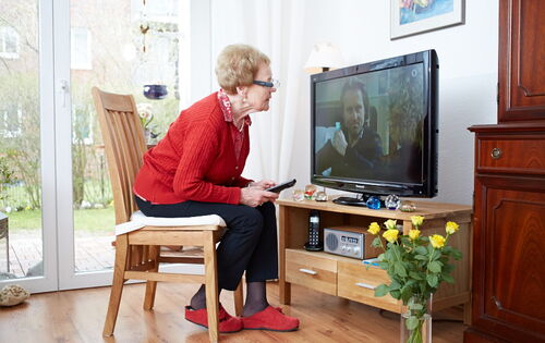 Eine ältere Frau in roter Strickjacke sitzt auf einem Stuhl direkt vor einem eingeschalteten Fernseher. In der Hand hält sie die Fernbedienung und beugt sich in Richtung des Bildschirms.